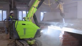 Зелёный робот‑демонтажник внутри здания: машина с навесным инструментом разрушает кирпичную стену, рядом оператор в каске.