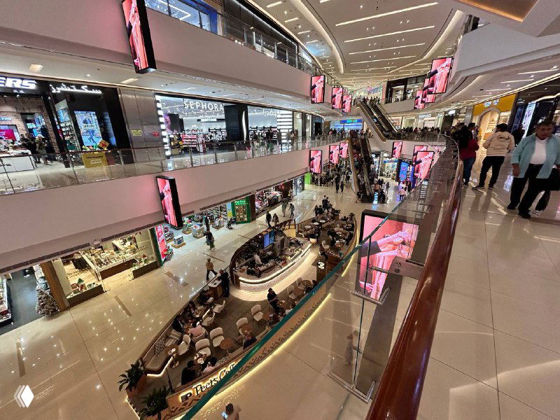 Многоуровневый торговый переход с магазинами по обеим сторонам, эскалаторы и людской поток внутри торгового центра.