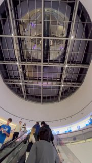 Вид снизу вверх на большой овальный атриум с куполом и верхними уровнями магазинов, посетители поднимаются по эскалатору.
