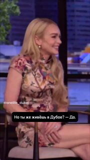 Женщина сидит в телевизионной студии в лёгком узорчатом платье; в кадре виден студийный фон и русская субтитровка "Но ты же живёшь в Дубае? — Да."