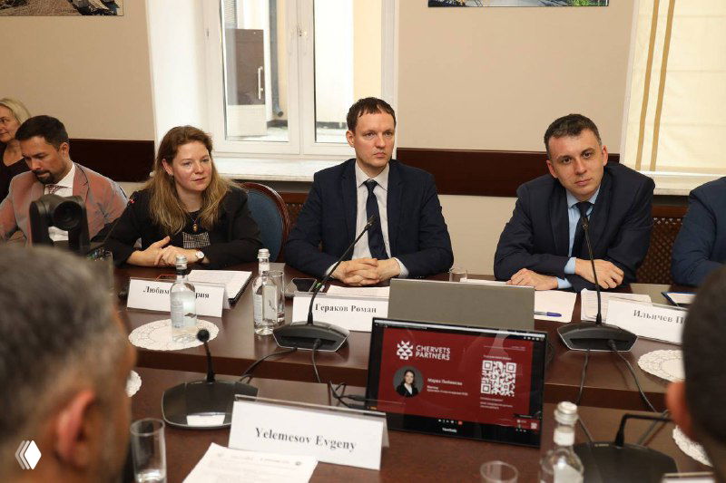 Группа участников круглого стола слушает выступления; на переднем плане таблички и планшеты с презентациями, видно деловую атмосферу встречи.