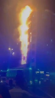 Ночной кадр: многоэтажный жилой дом в Дубае, фасад и балконы охвачены высоким пламенем, внизу видны силуэты людей и синий свет экстренных служб.