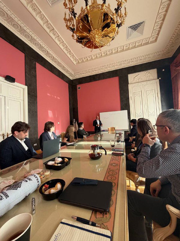 Переговорный стол в московском офисе: участники заседания, ноутбуки, чашки и закуски; в конце зала выступающий у доски — рабочая сессия.