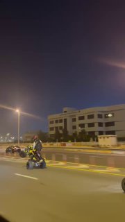 Группа гонщиков на квадроциклах едет по городской дороге в ночное время в Дубае; видны один водитель, бордюр, жилое здание и уличные фонари на фоне.