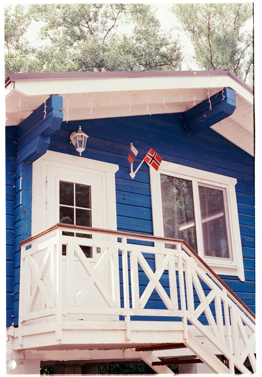 Скан кадра Kodak Vision3 250D: сине-белый дачный домик с маленьким балконом и флагами, спокойная цветопередача и тонкая зернистость плёнки.