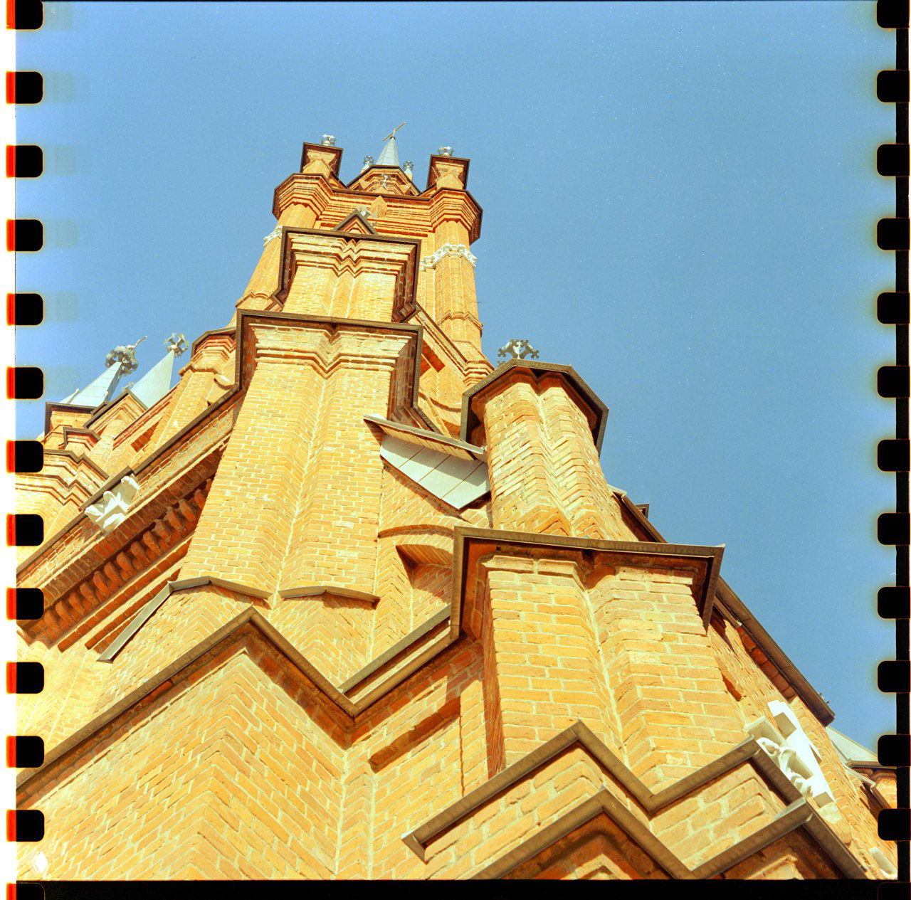 Вертикальный ракурс на высокую кирпичную башню костёла под голубым небом; видна текстура кирпича и краёв плёнки; Mamiya C220.