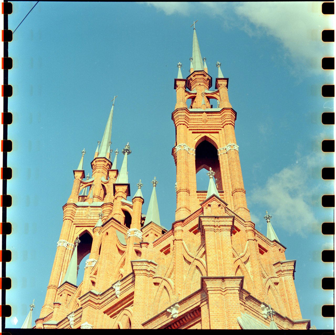 Силуэты шпилей и башен костёла в верхнем кадре, насыщенный голубой небосвод и контраст тёплого кирпича; цветная плёнка.