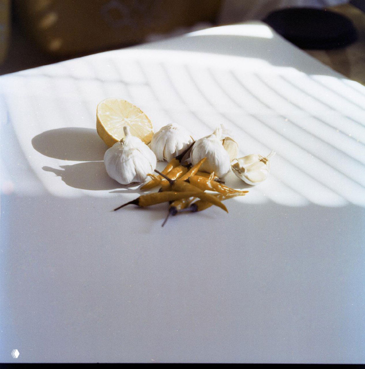 Лимон, головки чеснока и жёлтые острые перцы на белой поверхности с полосами тени от окна; плёночная гамма и мягкие тени.