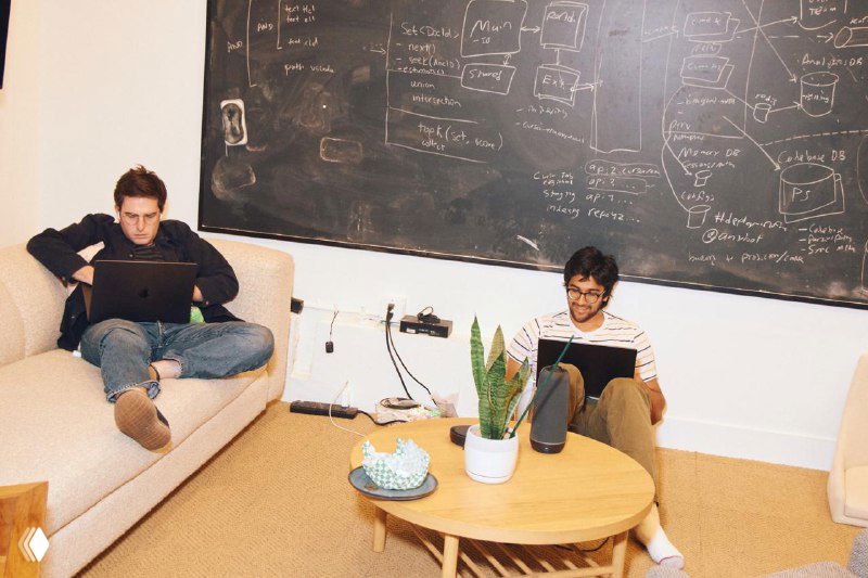 Зона для мозгового штурма в офисе Cursor: доска с диаграммами и заметками, двое сотрудников работают в уютной обстановке рядом с растениями.