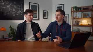 Два разработчика обсуждают код за столом с ноутбуком; на заднем плане доска с диаграммами и полки с книгами в офисе.