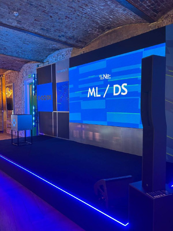 Фотография сцены конференции OZON E‑CODE 2025: большой LED-экран с надписью «ML/DS», сложная подсветка и профессиональная постановка.