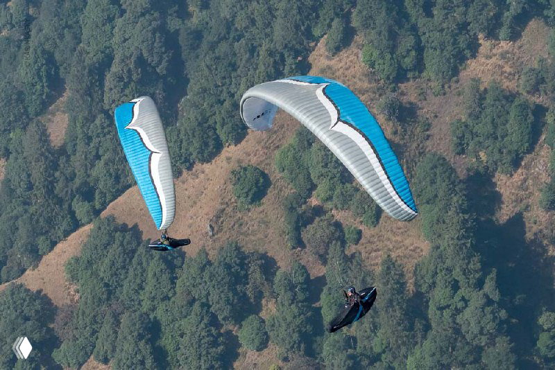 Два параплана в строю над зелёными хребтами; снимок подчёркивает форму крыла и взаимодействие с горным рельефом.