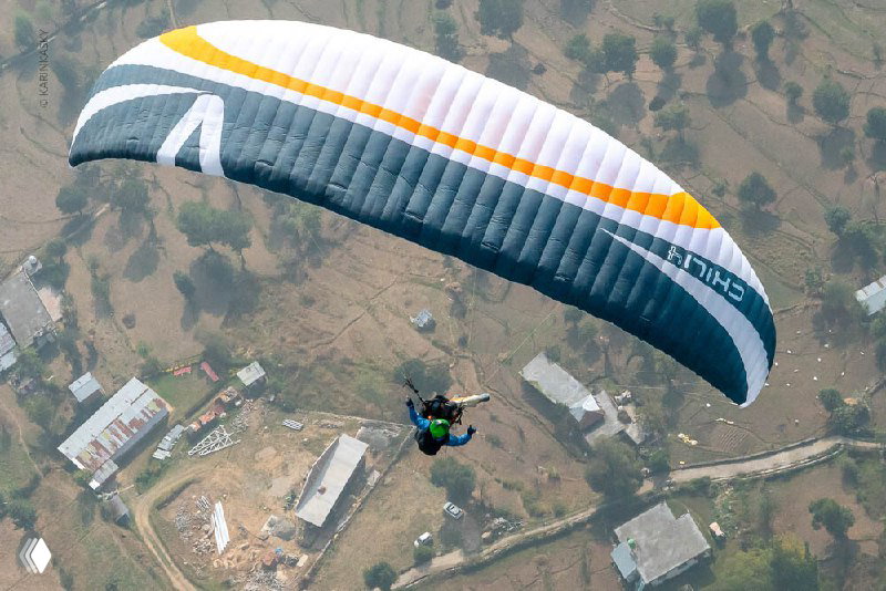 Крупный план бело-серого параплана над деревнями в долине; видна структура крыла и расположение домов внизу.