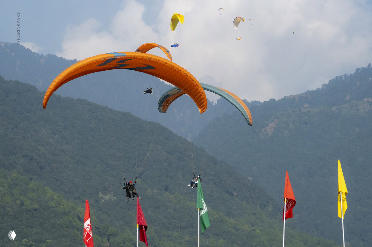 Несколько ярких куполов парапланов в небе над горным склоном и флагами; кадр показывает одновременный заход нескольких пилотов.