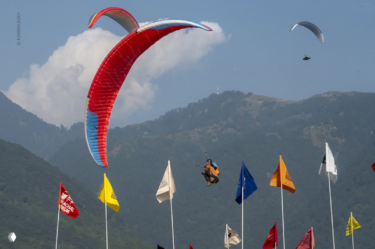 Красочный крупный параплан в небе над флагами и холмами; пилот на финальном заходе, видна динамика полёта и рельеф.