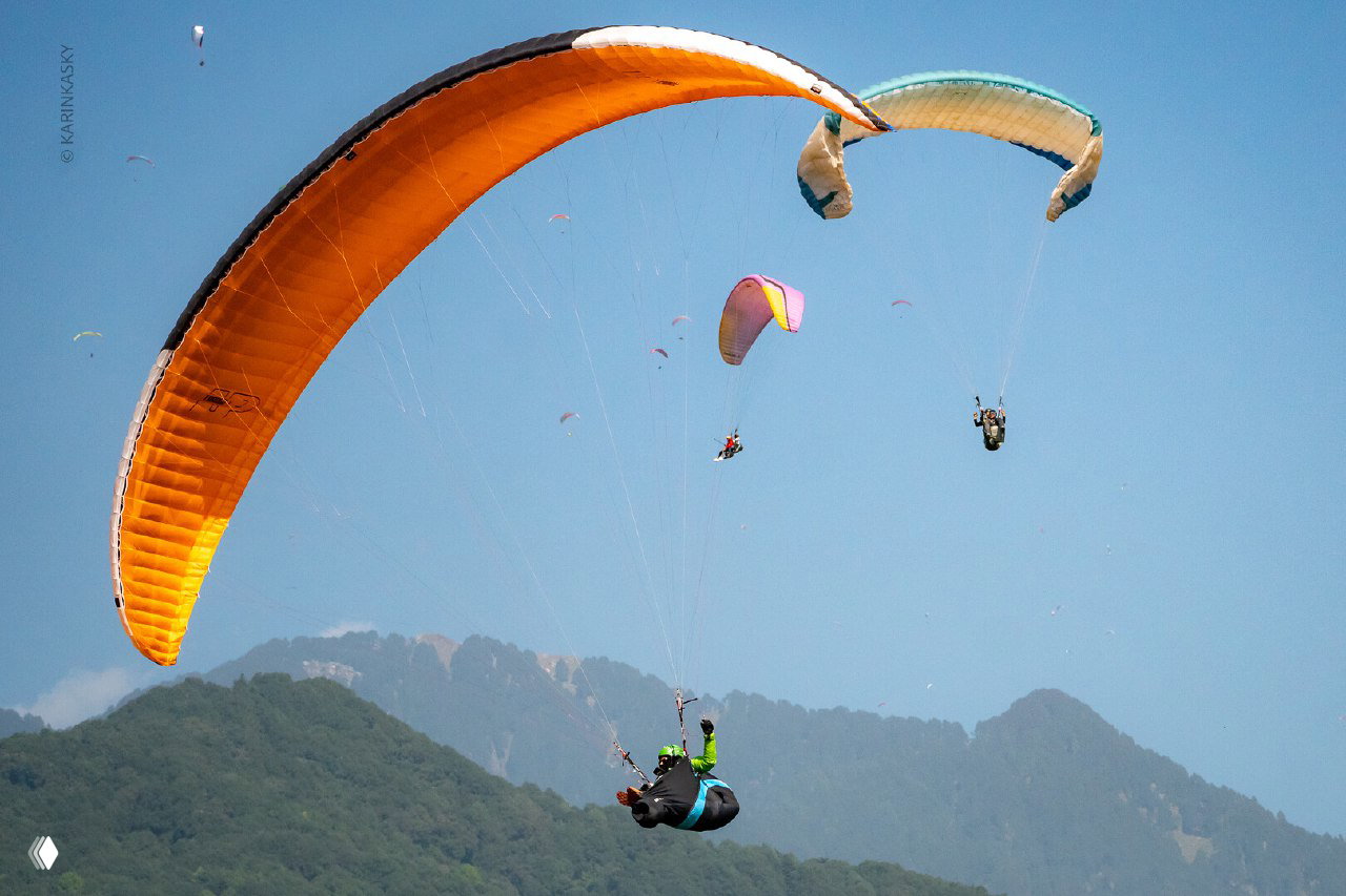 Оранжевый купол параплана в воздухе с несколькими другими крыльями вокруг; сцена иллюстрирует плотность воздушного трафика.
