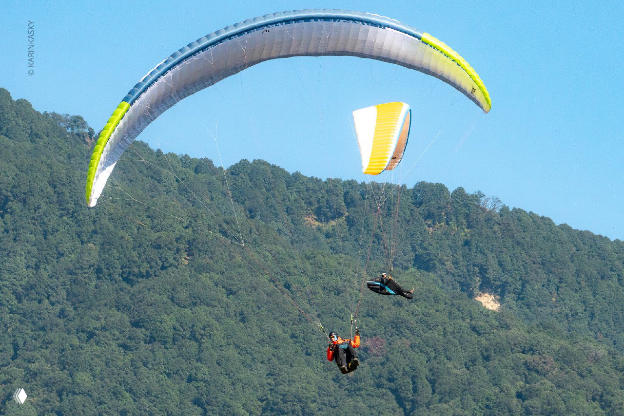 Голубой и бело-красный парапланы на фоне лесистого склона; несколько пилотов держат эшелон над рельефом.
