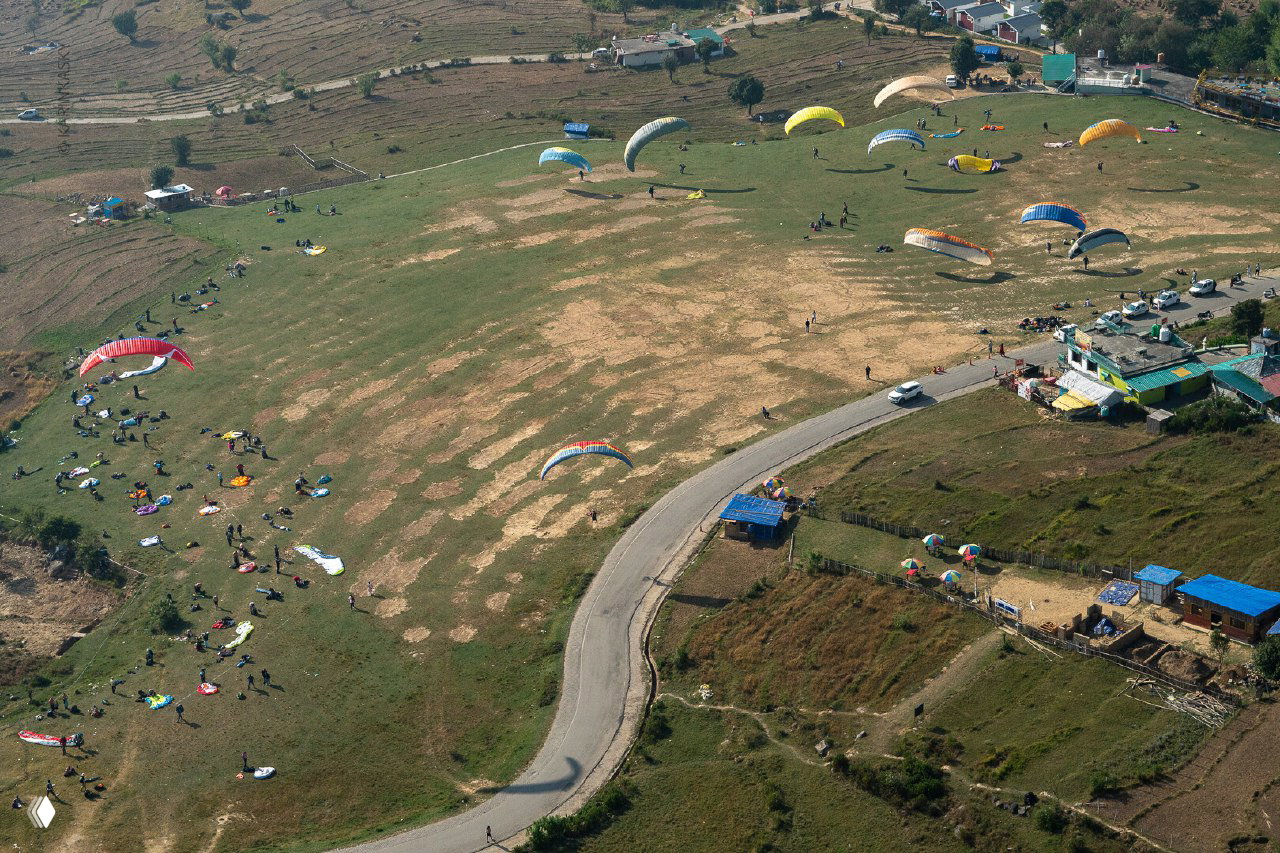 Вид сверху на посадочную поляну с расставленными парапланами и подходящими крыльями; на снимке видна распределённость пилотов по полю.