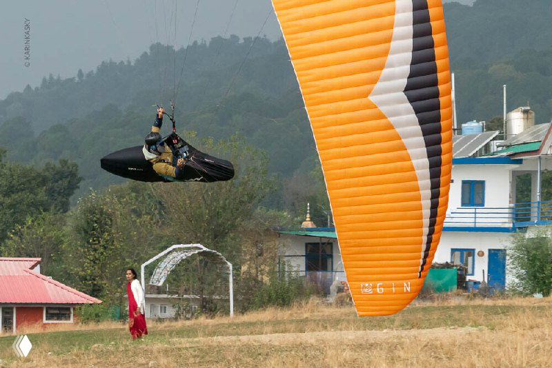 Парапланер делает низкий пролёт вблизи земли рядом с ярко-оранжевым крылом и домиками — подготовка к взлёту или посадке в индийской долине.