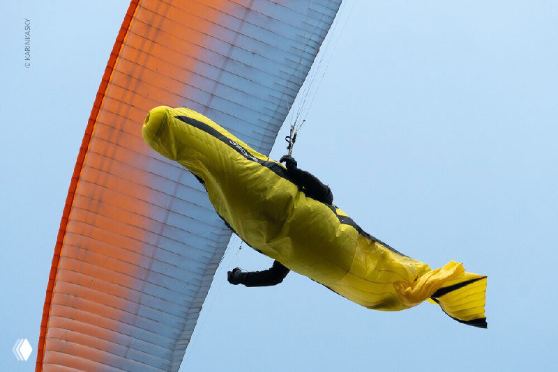 Крупный план пилота в жёлтом подвесе под парапланом, самолётность и экипировка видны на фоне небесной дистанции и канопей других пилотов.