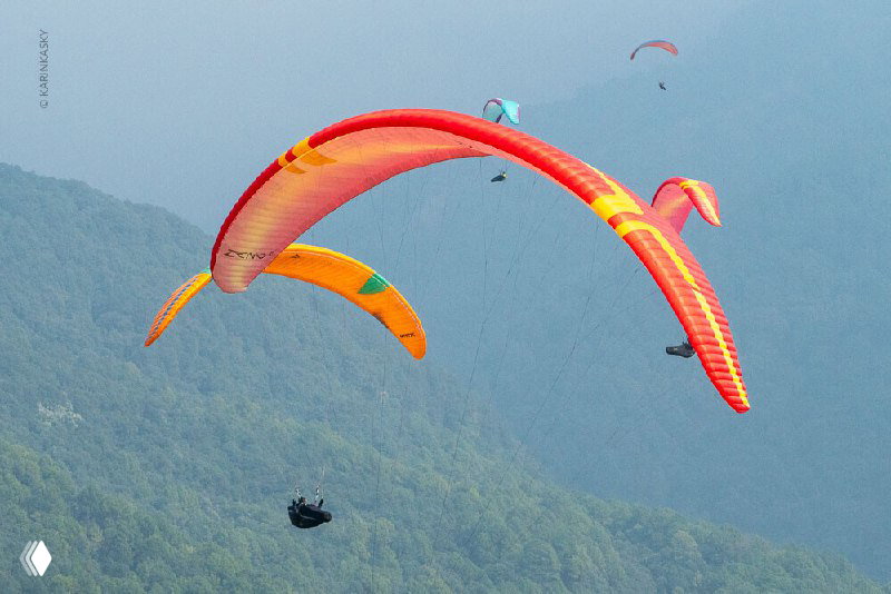 Несколько ярких парапланов оранжево-красной палитры на фоне зелёных склонов гор — сцена коллективного маршрута и обучения в Индии.