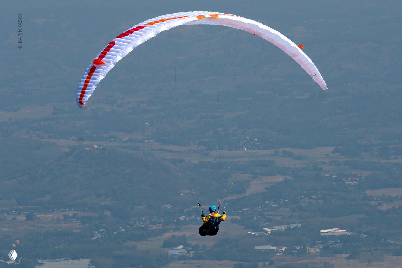 Парапланист в подвесной системе летит над равниной и холмами: одиночный кадр спортсмена во время дисциплины Tom Yum Cup 2024.