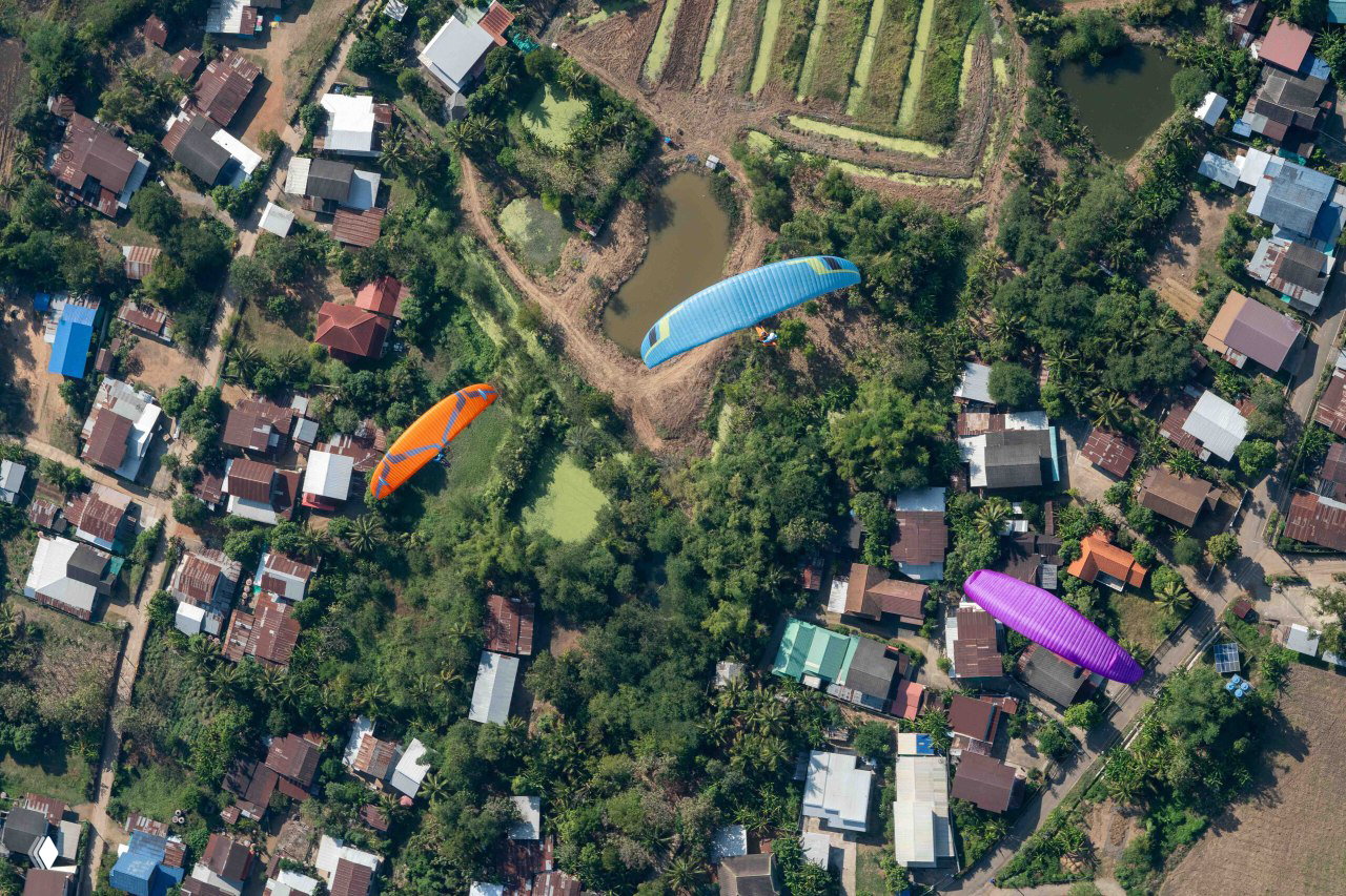 Несколько парапланов и разноцветные купола над сельской застройкой и заливными участками — панорамный вид этапа Tom Yum Cup 2024.