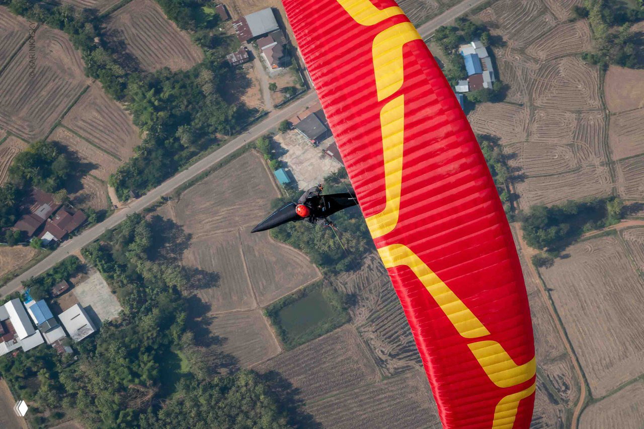 Красно-жёлтый параплан снят сверху, пилот в сидячем подвесе летит над полями и дорогой, отчётливый план местности под крылом.