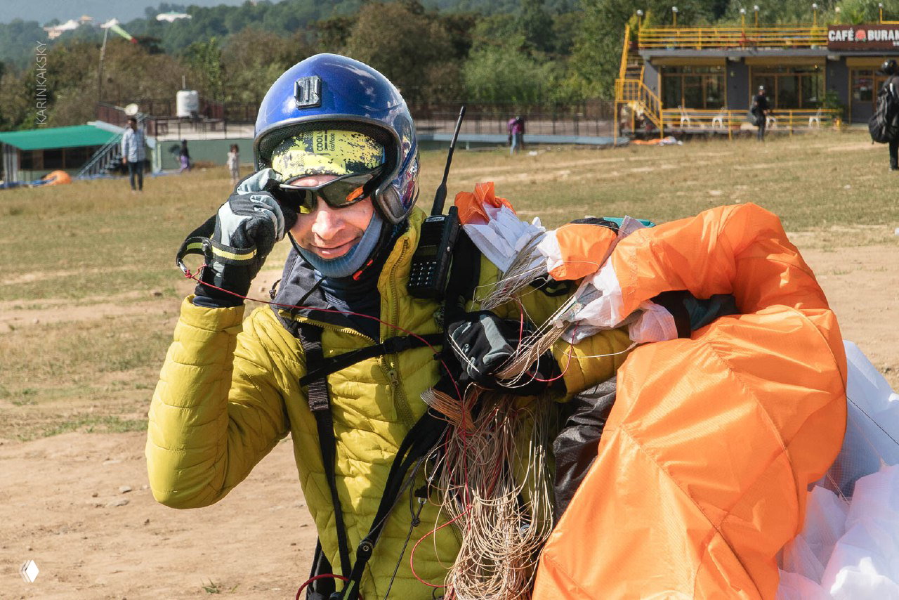 Парапланерист в шлеме и теплой куртке держит согнутый купол и стропы, проверяет снаряжение на старте на открытой площадке.