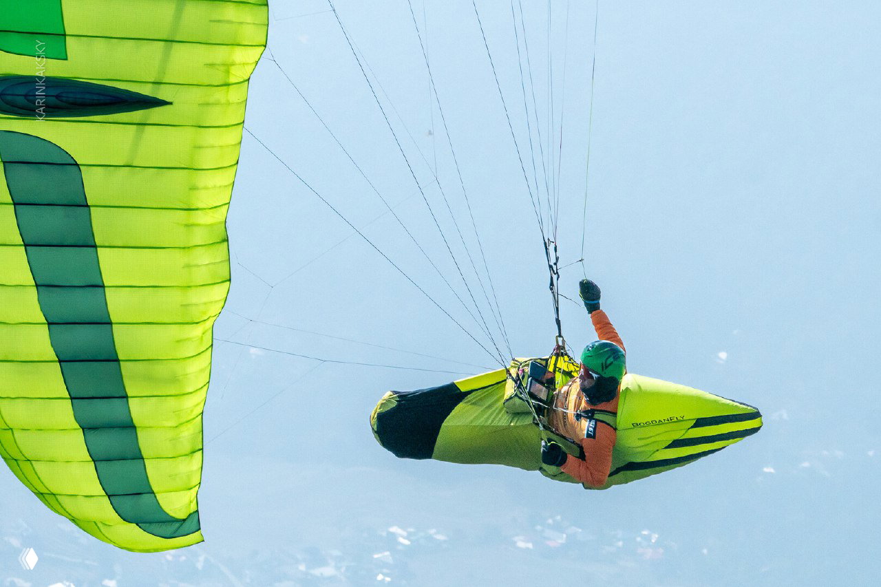 Парапланерист висит в подвесе под ярко-зеленым крылом, кадр с высоты показывает технику полета и положение тела пилота.