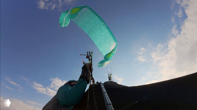 Пилот в подвесе смотрит вверх на бирюзовое крыло Artik 7P; видны тросы, стартовая конфигурация и небо над горизонтом, съёмка с позиции пилота.
