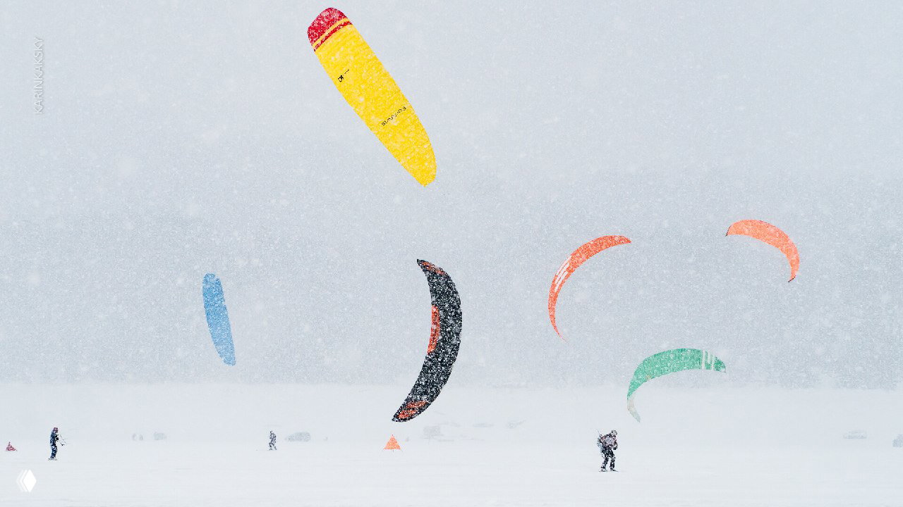 Несколько ярких кайтов в сильный снегопад над ровным льдом Красноярского моря: широкая панорама, маленькие фигуры людей на дистанции.