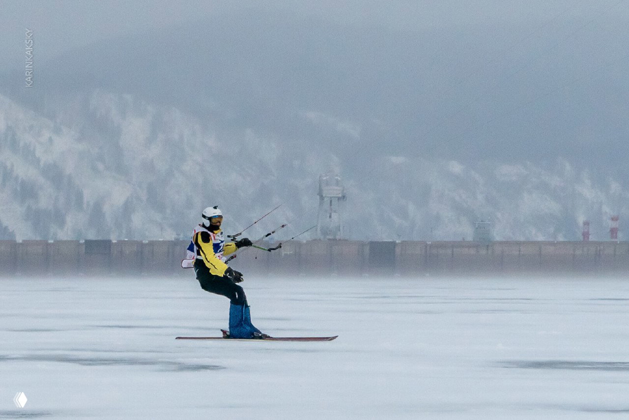 Кайтсёрфер на льду на фоне плотины и заснеженных холмов Красноярской ГЭС, спокойная зимняя сцена с акцентом на инфраструктуру.