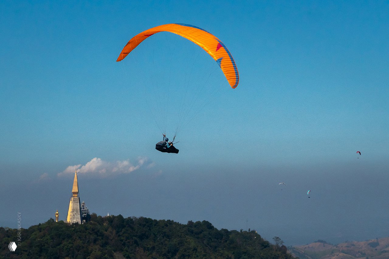 Оранжевый параплан в небе над холмом с видимой башней на вершине, чистое голубое небо и другие парапланы вдали формируют сцену полёта