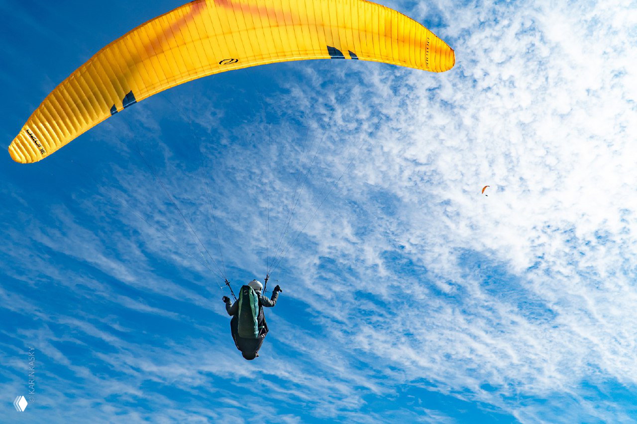 Вид снизу вверх на пилота под жёлтым парапланом на фоне узорчатых облаков, ощущение подъёма и пространства в небе