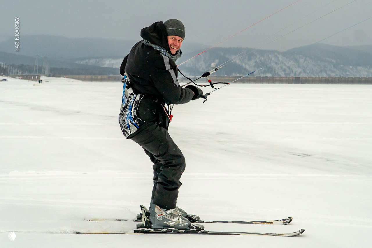 Улыбающийся спортсмен на лыжах управляет кайт-управлением на льду Красморе; видно снаряжение для сноукайтинга и удалённые холмы — сезон 2025.
