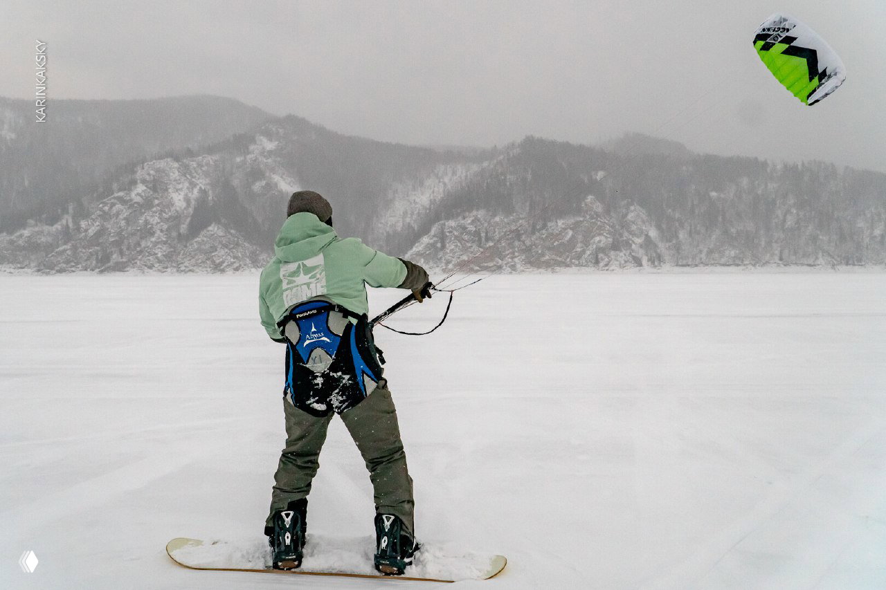 Кайтсерфер на сноуборде стоит на льду Красморе, тянет линии кайта на фоне скалистых гор и серого неба — типичная зимняя сессия 2025.