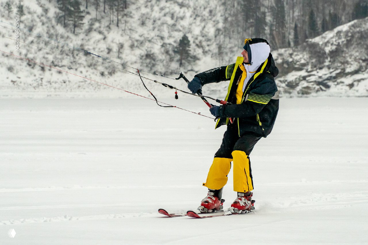 Спортсмен в яркой куртке и жёлтых штанах катается на лыжах с кайтом по ровному льду; видны линии управления и снежные склоны Красморе — сезон 2025.