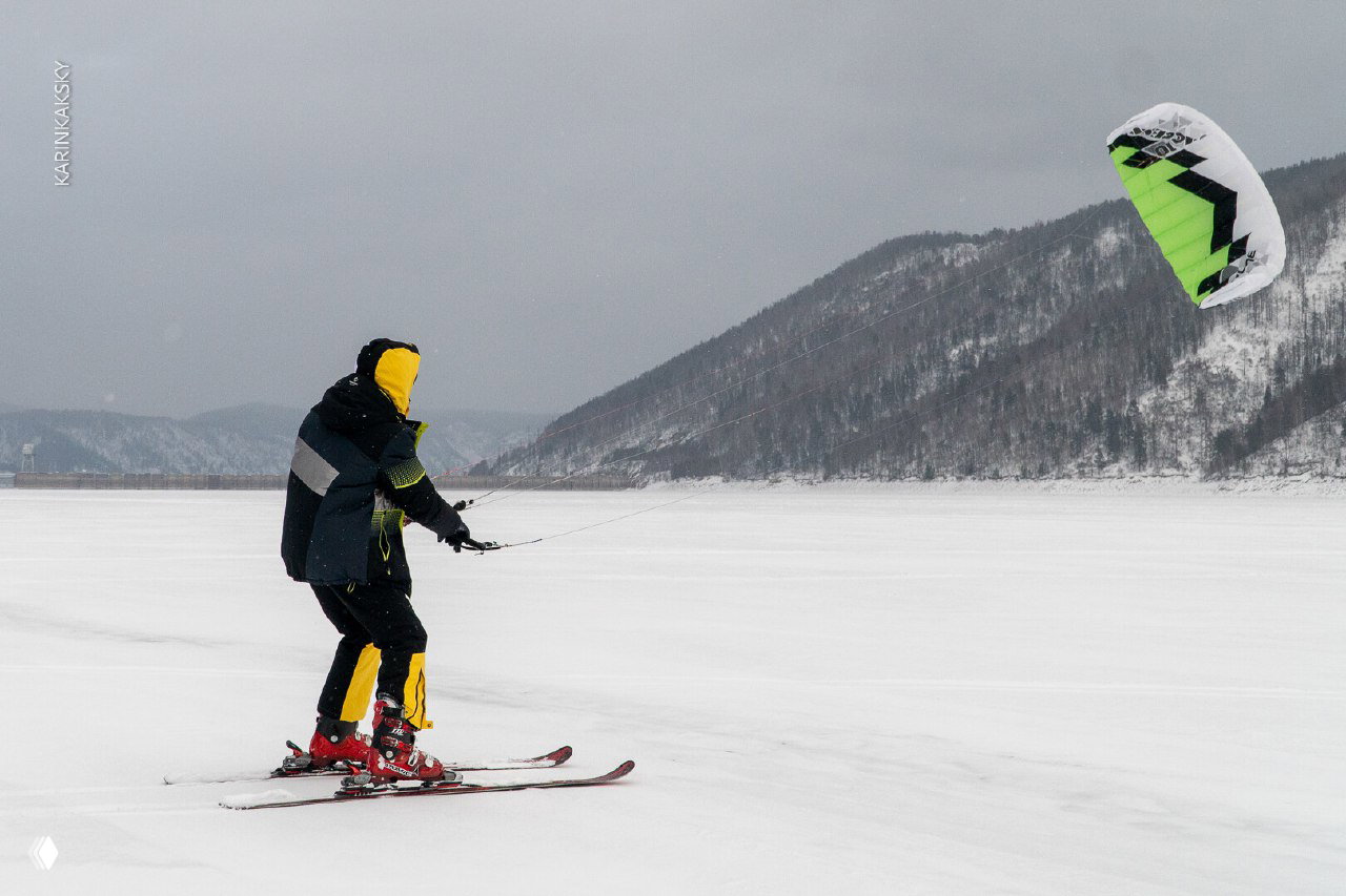 Кайт в небе над заснеженным озером и человек на лыжах управляет кайтом на фоне дальних гор; широкая панорама льда и пейзажа Красморе — 2025.