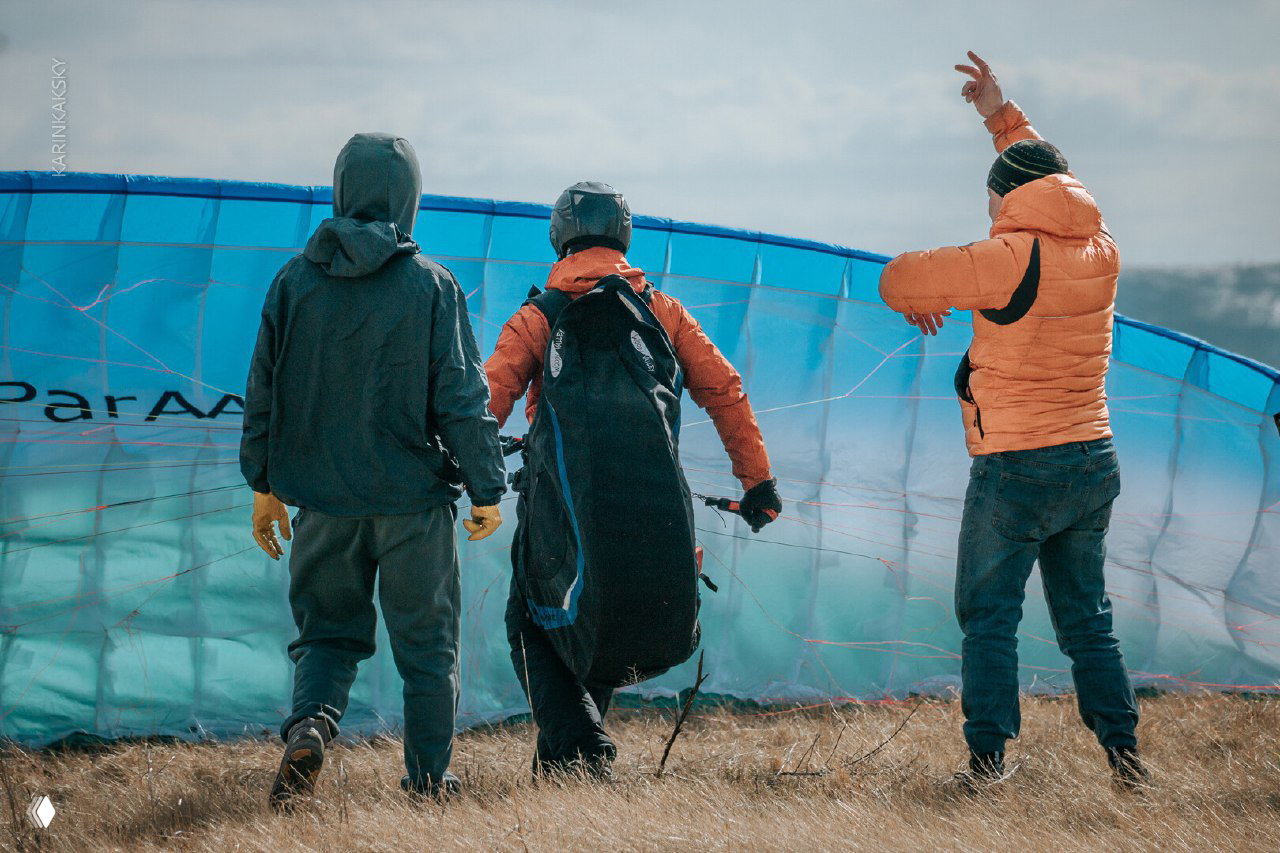 Трое людей готовят параплан к запуску на травянистом склоне: инструктор и ученики в шлемах и куртках расправляют купол и стропы.