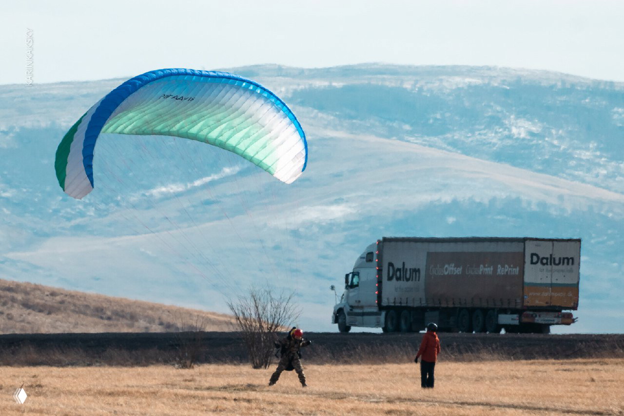 Параплан бело-зелёного цвета поднимается на фоне холмов; на переднем плане человек с парашютом, на дороге видна грузовая фура вдали.