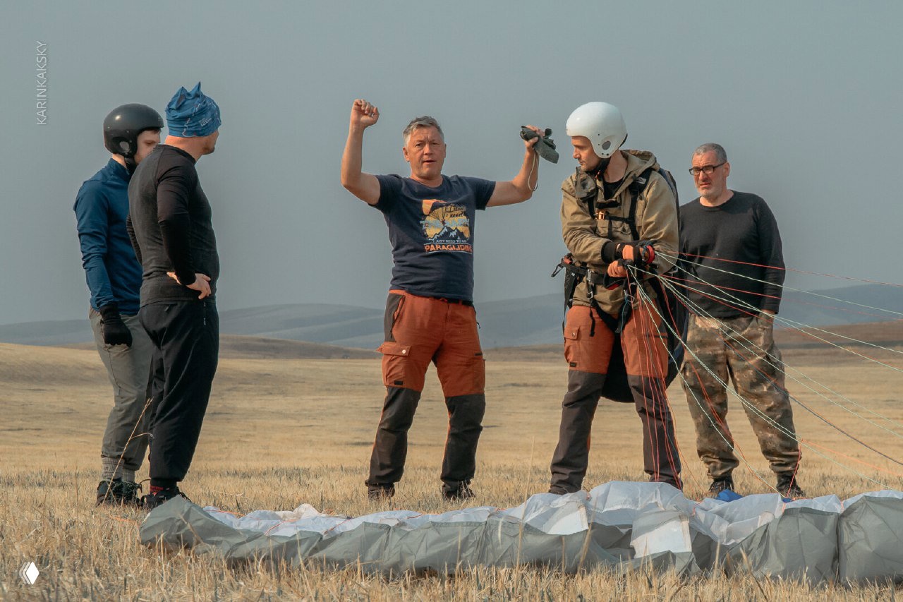 Группа пилотов на поле: инструктор объясняет технику перед полётом, видна расправленная ткань купола и подготовка снаряжения.