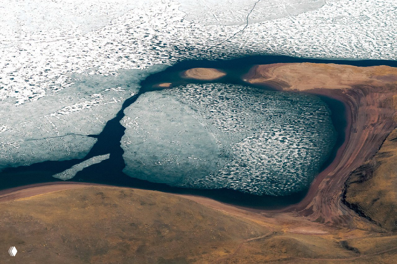 Аэрофотоснимок весеннего пейзажа: остатки льда на воде, изрезанные береговые контуры и тёплые земляные тона побережья с естественной текстурой.