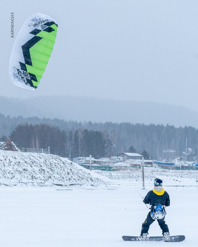 Райдер на лыжах едет по льду Красморе у береговой линии: зелёный кайт в воздухе, рядом видны дома и лесная полоса на горизонте за берегом.