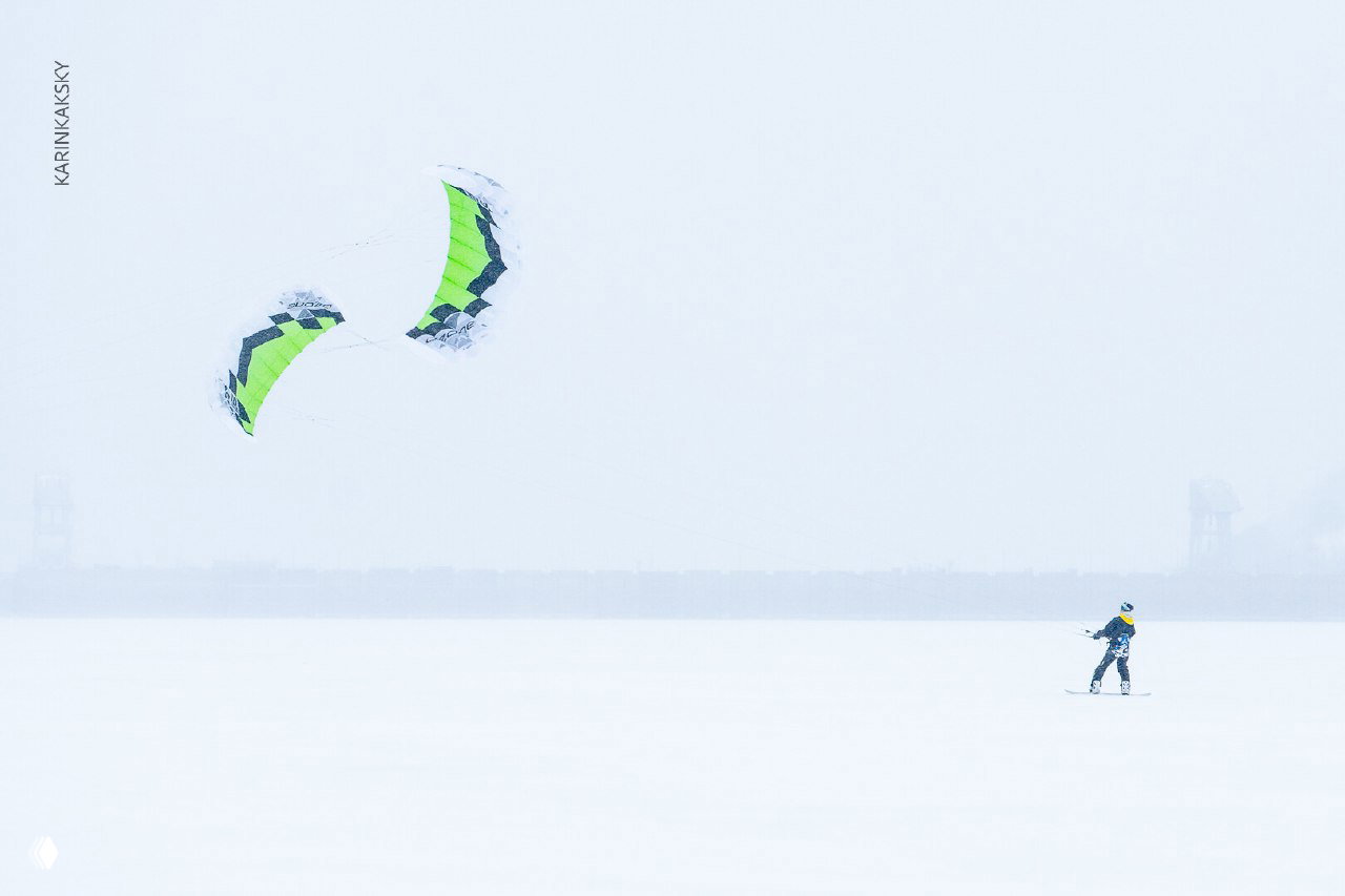 Широкий минималистичный кадр Красморе: маленькая фигура райдера и крупный зелёный кайт над пустым белым пространством льда и туманной линией горизонта.