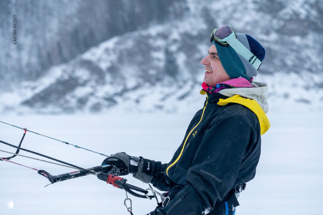 Портрет райдера в шлеме и очках на Красморе: улыбающийся человек управляет кайтом, на фоне заснеженный склон и лес, снег слегка падает.