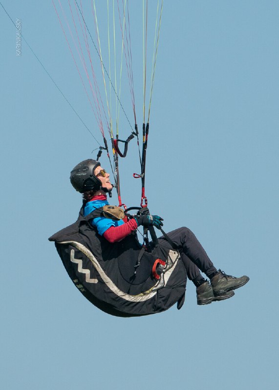 Парапланерист в подвеске висит в воздухе на фоне чистого неба — видна конструкция подвески и управление страховочными линиями.