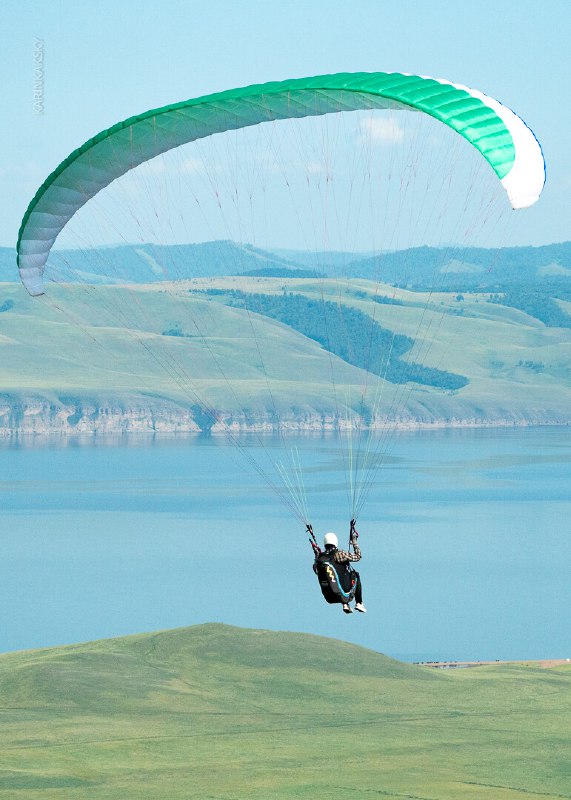 Парапланерист летит над холмами и озером на светлом параплане, видно ландшафт и спокойные условия для маршрутов.
