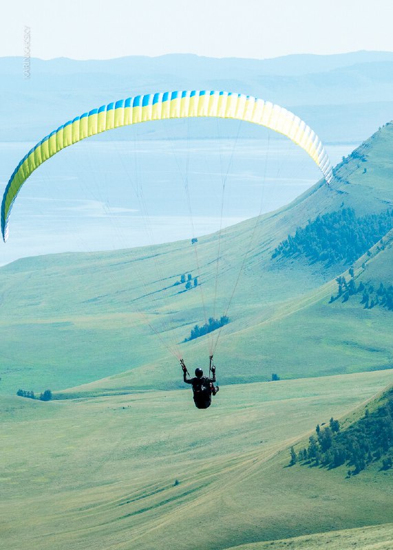 Один пилот на фоне берегового ландшафта и озера держится в воздухе на двухцветном параплане — видна дистанция и простор полёта.