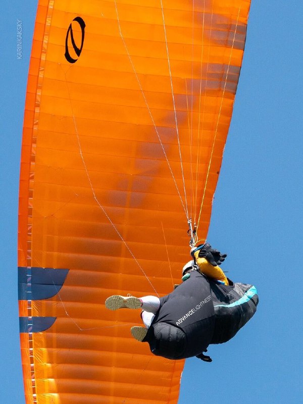 Крупный план пилота в подвеске и ярко‑оранжевого крыла, кадр снизу; видны стропы, посадочная поза и детали управления в полёте.